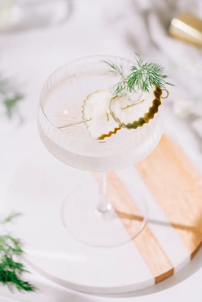 An overhead shot of a martini glass filled with clear liquid on a tablescape with two pickle slices and a dill frond as a garnish.