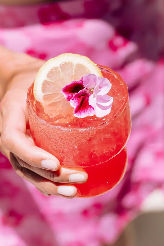 A sparkly pink drink with an edible flower and lemon