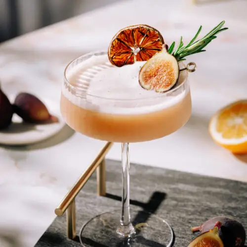 A whiskey cocktail in a coupe glass garnished with fig, dehydrated lemon, and rosemary sprig on a black tray table.