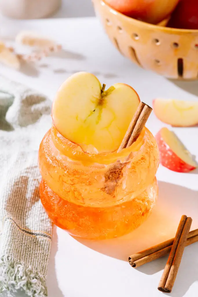 Apple-rol Spritz drink in a glass with an apple slice and cinnamon stick as garnish.