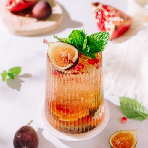 A fall cocktail with reddish liquid, topped with figs, mint, and pomegranate arils.