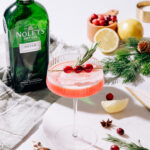 Cranberry Olio Gin Sour in a martini glass, garnished with cranberries and a rosemary sprig. The NOLET's green bottle is in the background.