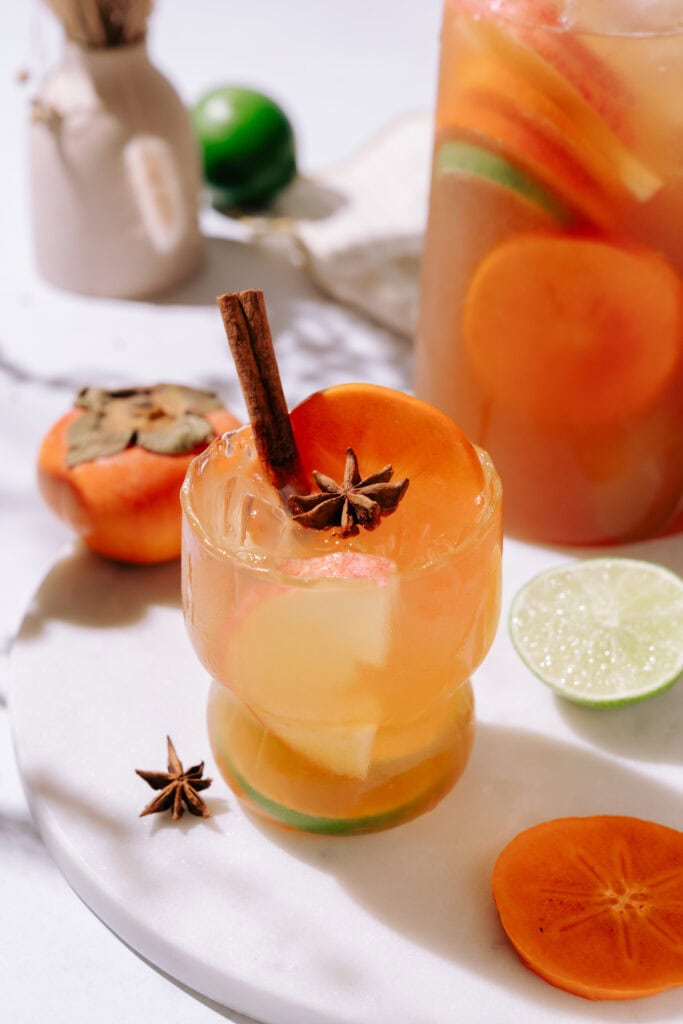 A cocktail glass with Moscow Mule Thanksgiving Sangria. With a cinnamon stick and star anise as garnish. Lime and persimmon are on table behind it and a pitcher of sangria is visible.