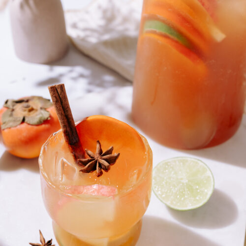 A cocktail glass with orange liquid, a cinnamon stick and star anise as garnish. Lime and persimmon are on table behind it and a pitcher of sangria is visible.