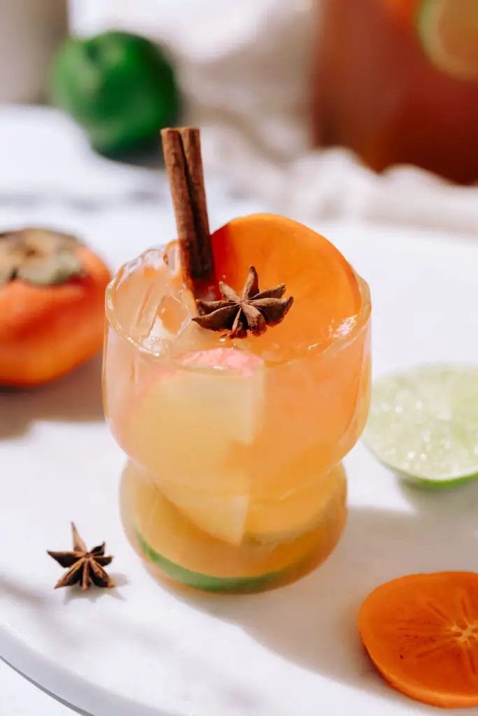 A cocktail glass with orange liquid, a cinnamon stick and star anise as garnish. Lime and persimmon are on table behind it.