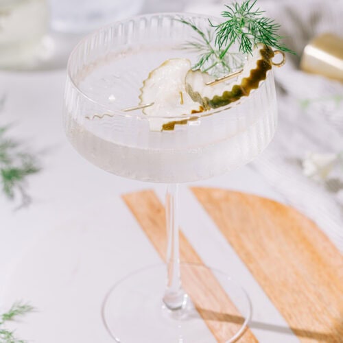 An overhead shot of a martini glass filled with clear liquid on a tablescape with two pickle slices and a dill frond as a garnish.