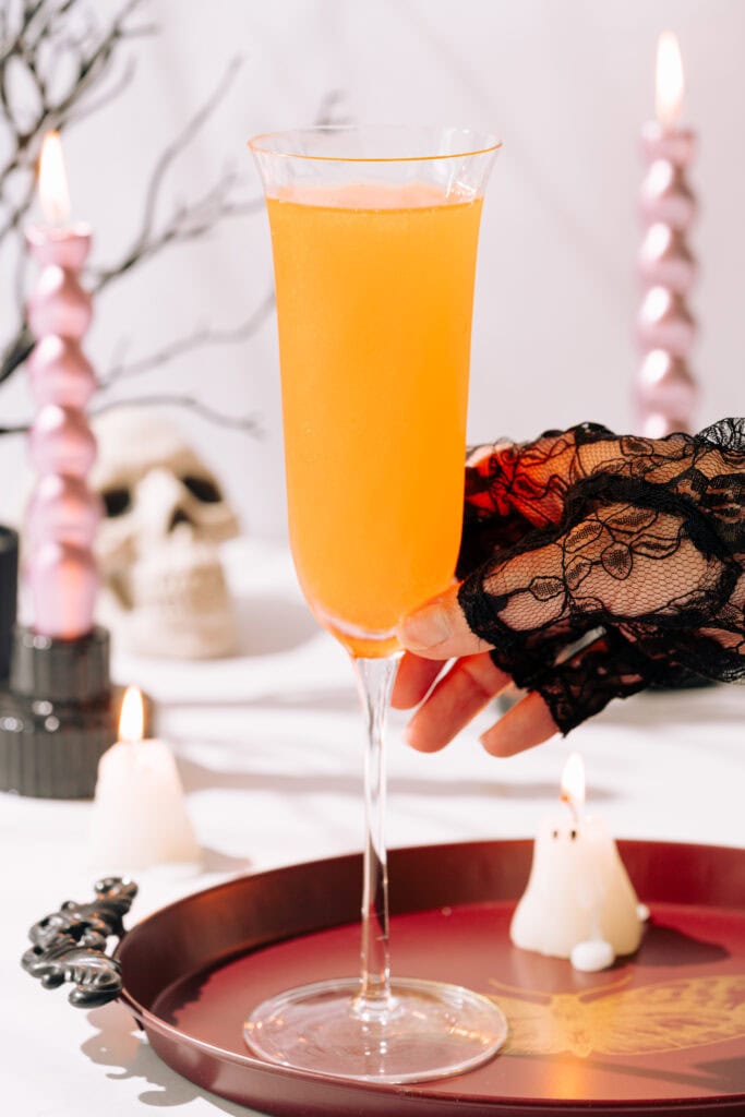 A hand in a black lace glove is reaching for a mimosa in a champagne flute on a burgundy tray