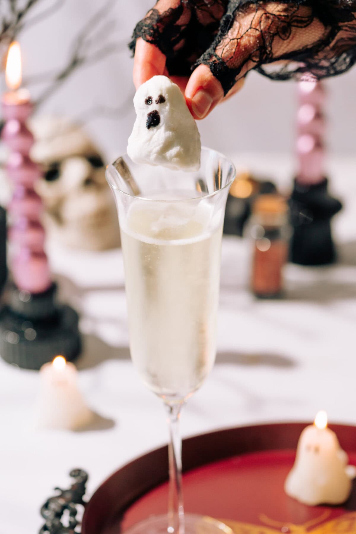 A hand wearing a black lace glove drops a ghost glitter bomb into a champagne flute filled with champagne.