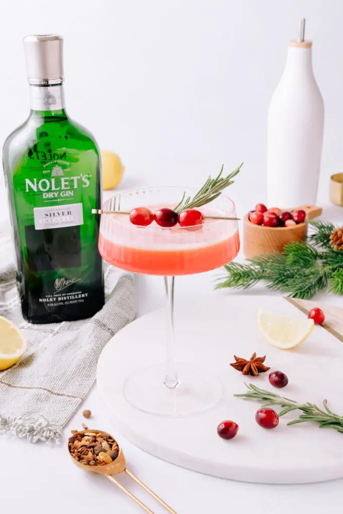 Cranberry Olio Gin Sour in a martini glass, garnished with cranberries and a rosemary sprig. The NOLET's green bottle is in the background. 