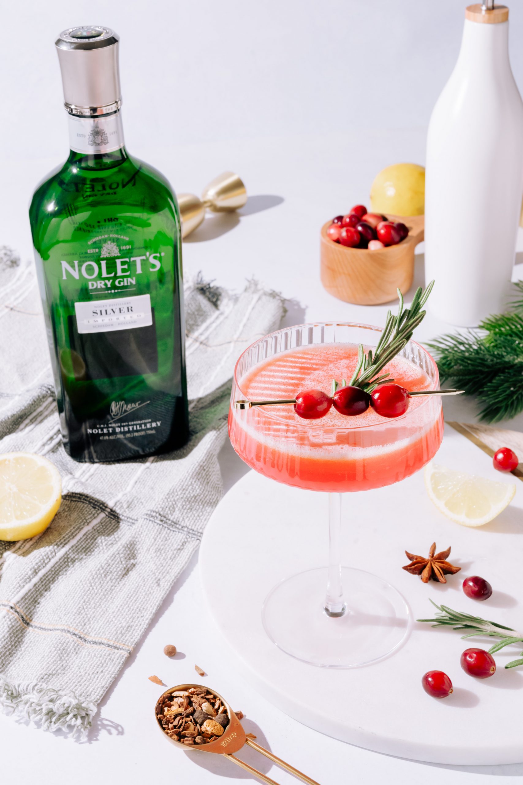 Cranberry Olio Gin Sour in a martini glass, garnished with cranberries and a rosemary sprig. The NOLET's green bottle is in the background.