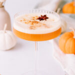 A coupe glass with orange liquid is topped with foam, a sprinkle of spice, and star anise. Cinnamon and pumpkins are on the table.