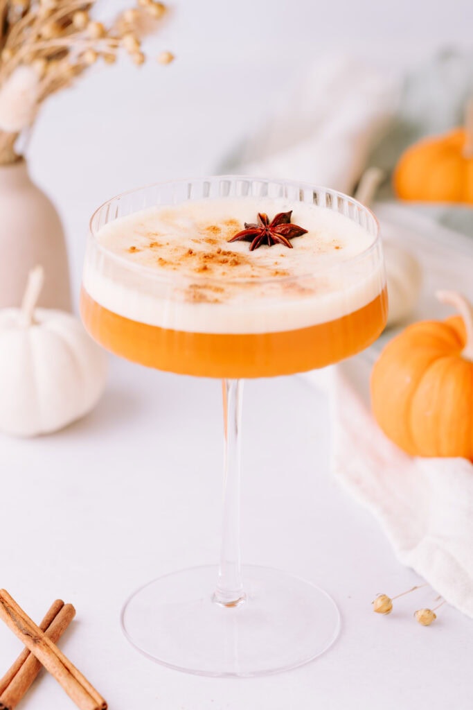 A coupe glass with orange liquid is topped with foam, a sprinkle of spice, and star anise. Cinnamon and pumpkins are on the table.
