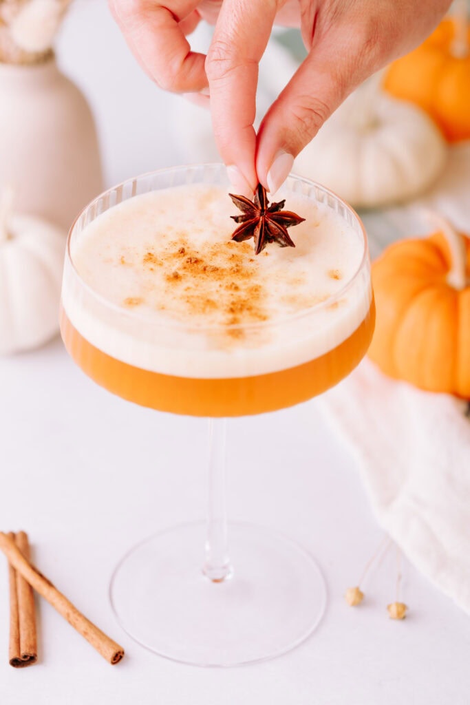 A coupe glass with orange liquid is topped with foam, a sprinkle of spice, and star anise. Cinnamon and pumpkins are on the table.