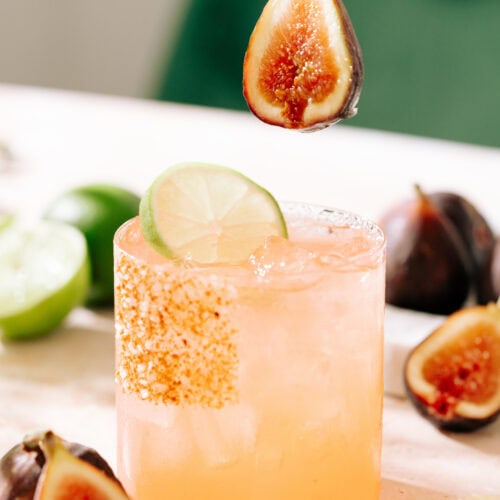 maple fig margarita cocktail with a fresh fig being placed in the glass. The rim is coated with sugar, salt, and cinnamon.