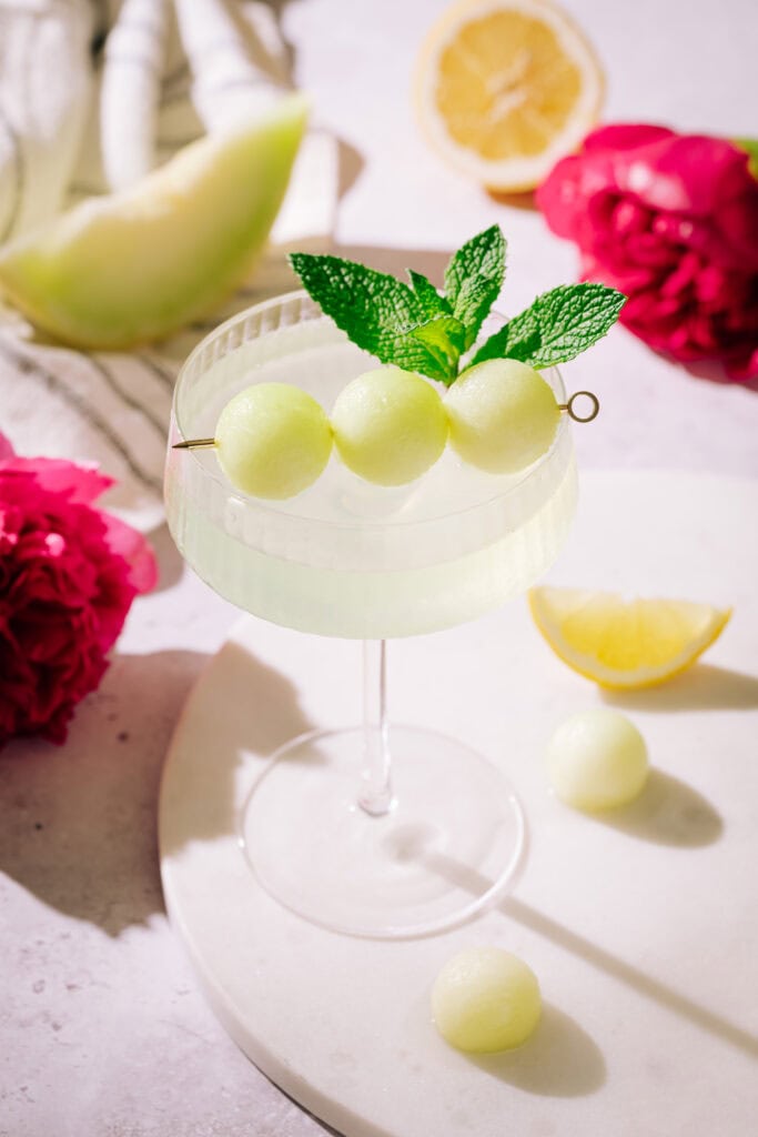 Honeydew appletini garnished with 3 melon balls on a tray surrounded by fruit and flowers