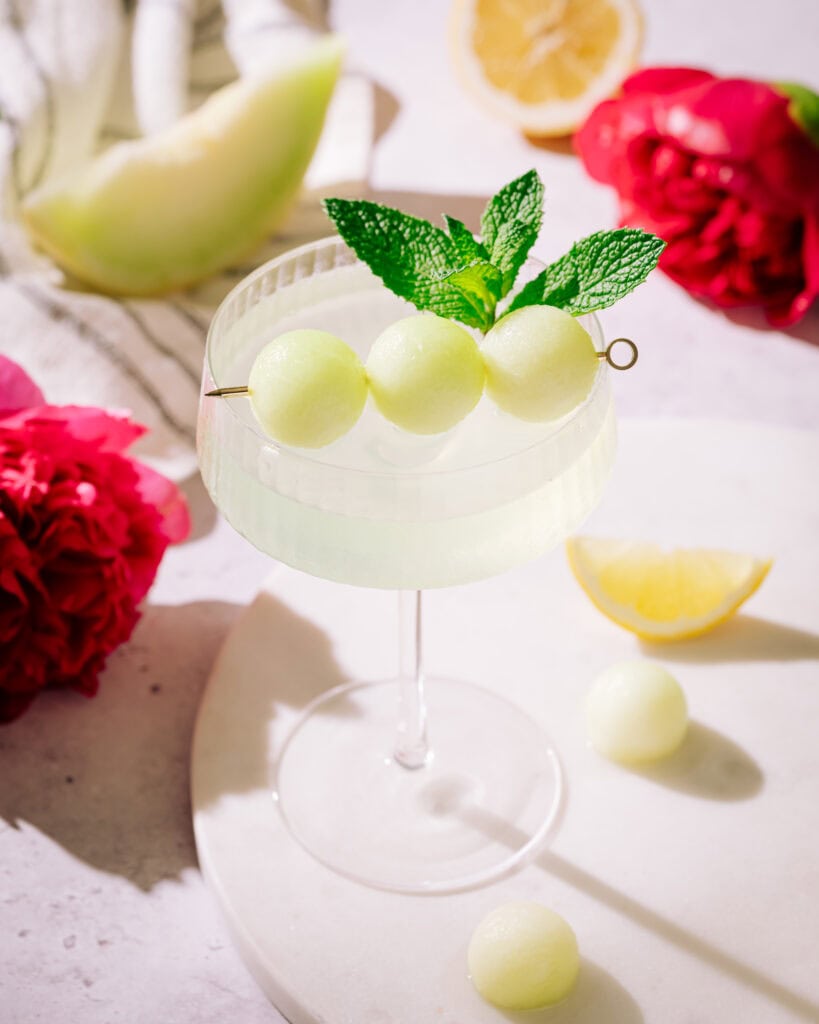 Honeydew Appletini cocktail on board, garnished with 3 melon balls on a pick and a mint sprig