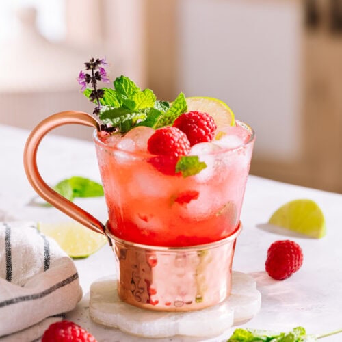 Raspberry moscow mule in glass and copper mug on coaster garnished with raspberries, lime and mint