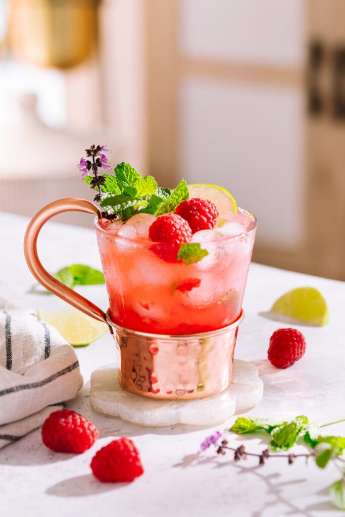 Raspberry moscow mule in glass and copper mug on coaster garnished with raspberries, lime and mint