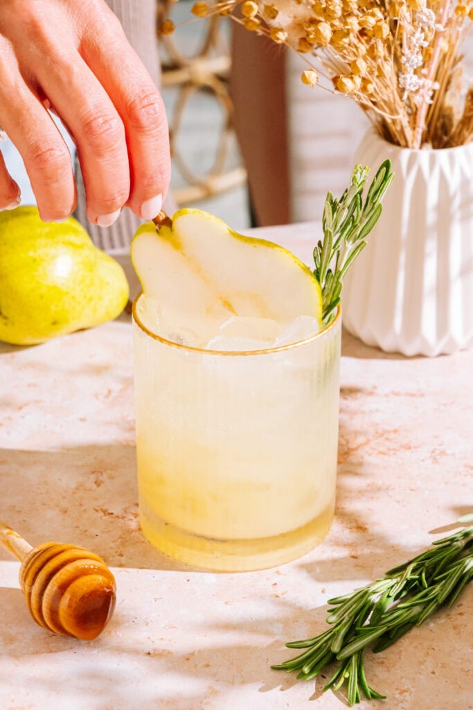 a hand placing a slice of pear into the rosemary pear mocktail on the table. A sprig of rosemary and a wooden honey dipper is also on the table.