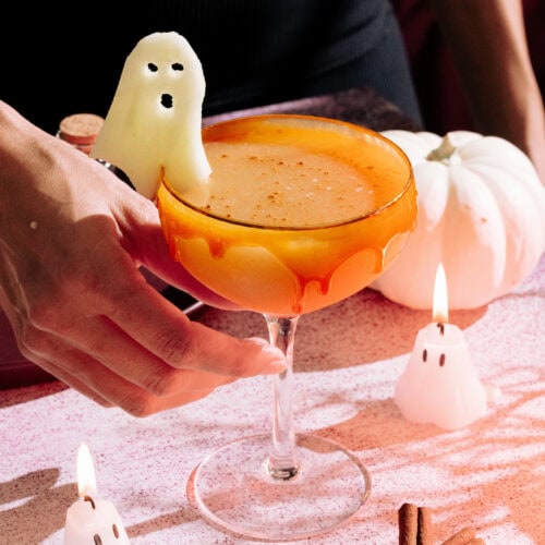 a hand reaching for the Harvest Apple (Apple Cider Whiskey Cocktail) with a caramel rim, ghost apple garnish on a table with mini ghost candles and a white pumpkin.