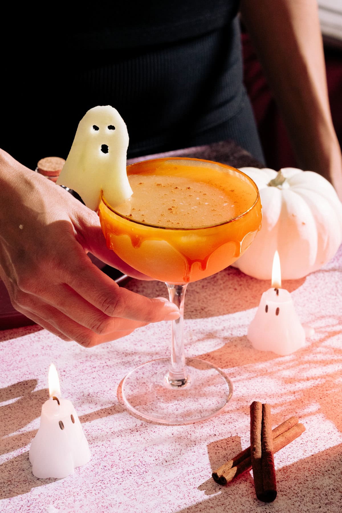 a hand reaching for the Harvest Apple (Apple Cider Whiskey Cocktail) with a caramel rim, ghost apple garnish on a table with mini ghost candles and a white pumpkin.