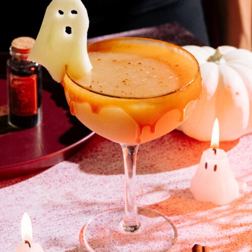 The Harvest Apple (Apple Cider Whiskey Cocktail) with a caramel rim, ghost apple garnish on a table with mini ghost candles and a white pumpkin.