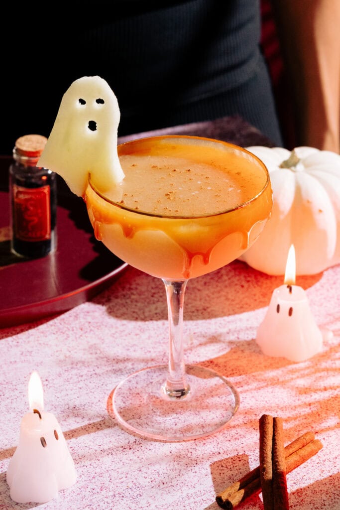 The Harvest Apple (Apple Cider Whiskey Cocktail) with a caramel rim, ghost apple garnish on a table with mini ghost candles and a white pumpkin.