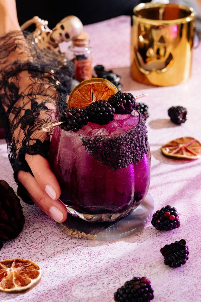 a hand wearing a lace glove reaches for the midnight margarita. it is rimmed with black salt and garnished with blackberries and a dehydrated lime