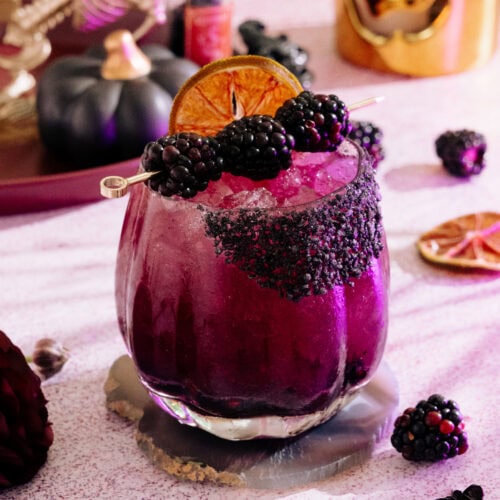 a blackberry margarita with a dark purple hue, rimmed with a black salt rim, and garnished with blackberries and dehydrated lime