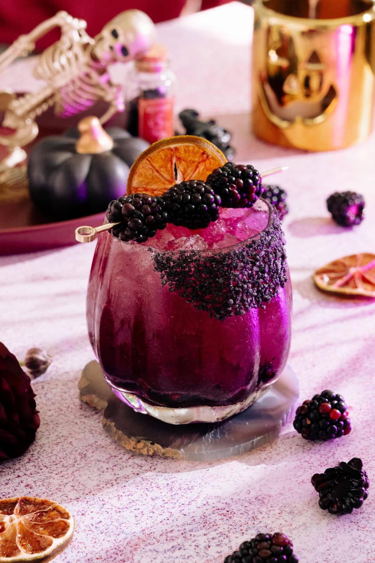 a blackberry margarita with a dark purple hue, rimmed with a black salt rim, and garnished with blackberries and dehydrated lime