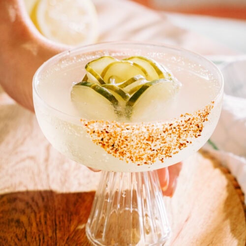 a hand reaches for a pickle mimosa with a pickle ice cube and tajin rim