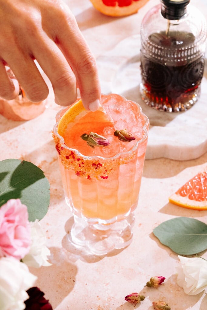 Empress elderflower rose gin cocktail with a brown sugar, dried rose, and grapefruit zest rim. Two dried rose buds garnish the cocktail, and a hand is placing a grapefruit slice in the cocktail as another garnish. A bottle of brown sugar syrup is in the background on the table. 