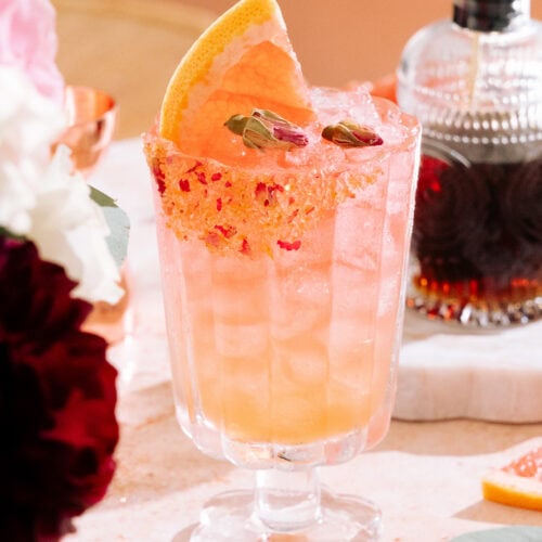 Empress elderflower rose gin cocktail with a brown sugar, dried rose, and grapefruit zest rim. Rose buds and a fresh grapefruit slice garnish the cocktail. A bottle of brown sugar syrup is in the background on the table.