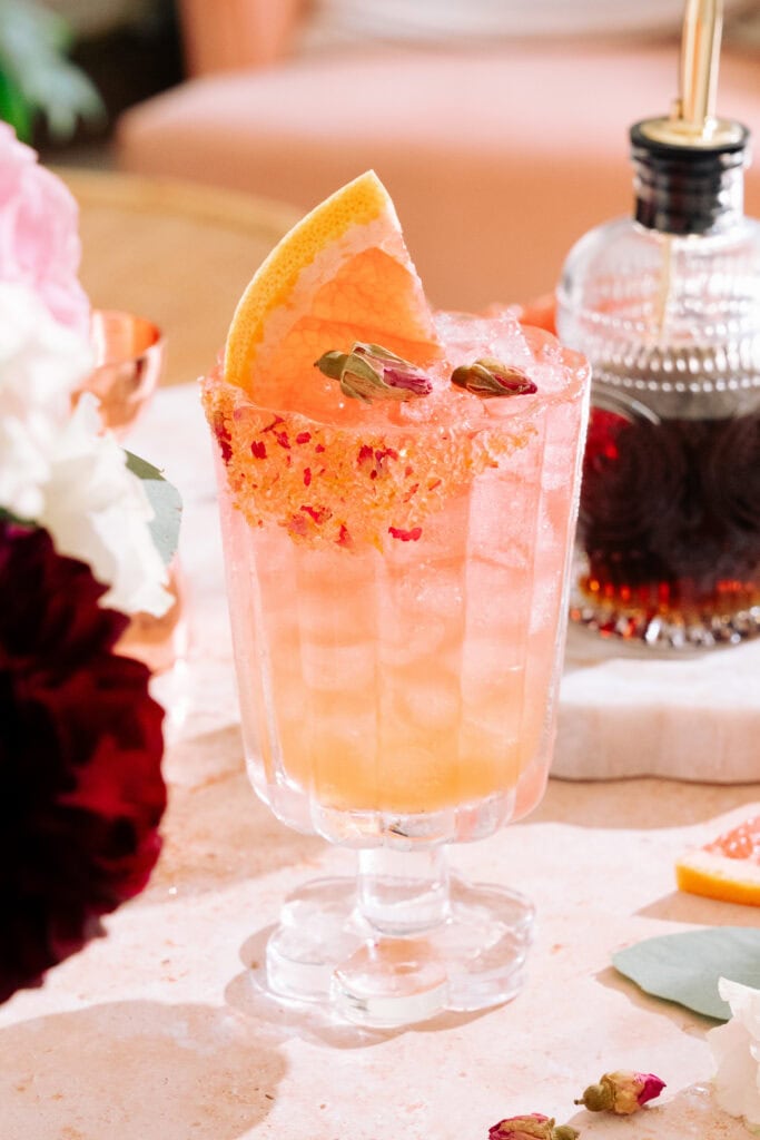 Empress elderflower rose gin cocktail with a brown sugar, dried rose, and grapefruit zest rim. Two dried rose buds and a fresh grapefruit slice garnish the cocktail. A bottle of brown sugar syrup is in the background on the table.