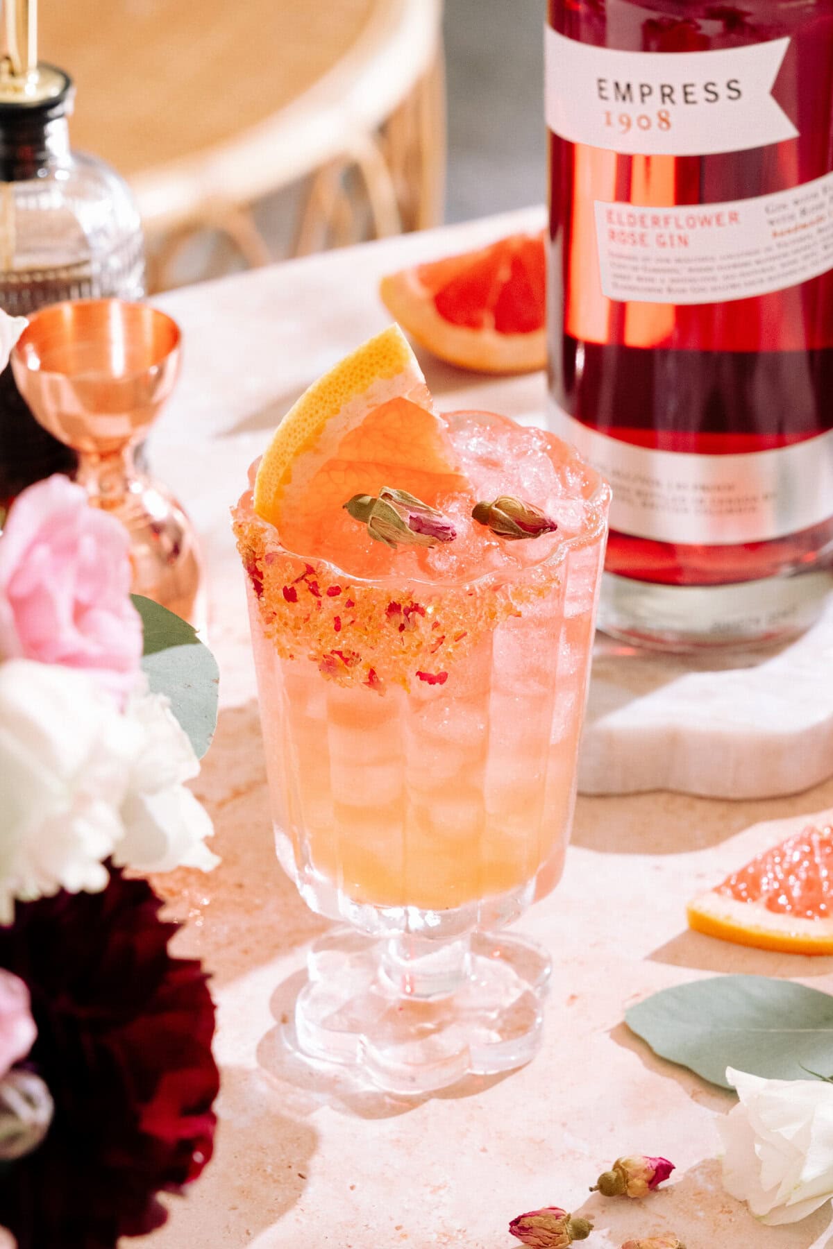 Empress elderflower rose gin cocktail with a brown sugar, dried rose, and grapefruit zest rim. Garnished with a grapefruit slice and two dried rose buds. A bottle of Empress elderflower rose gin is in the background.