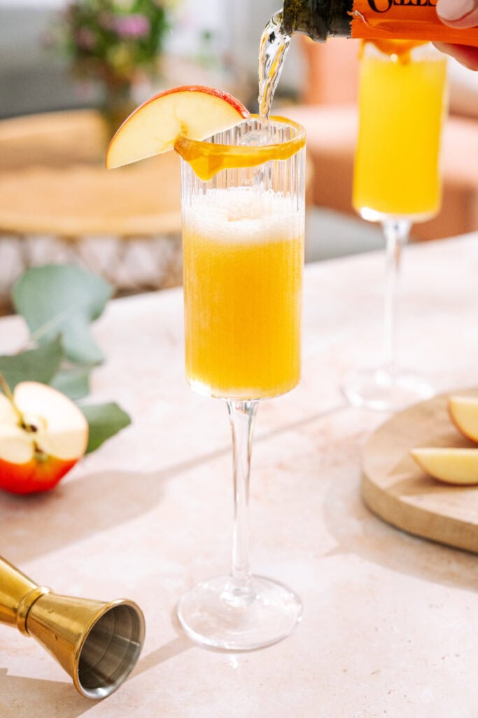 Prosecco pouring into Caramel apple mimosa with caramel rim, garnished with apple slice.