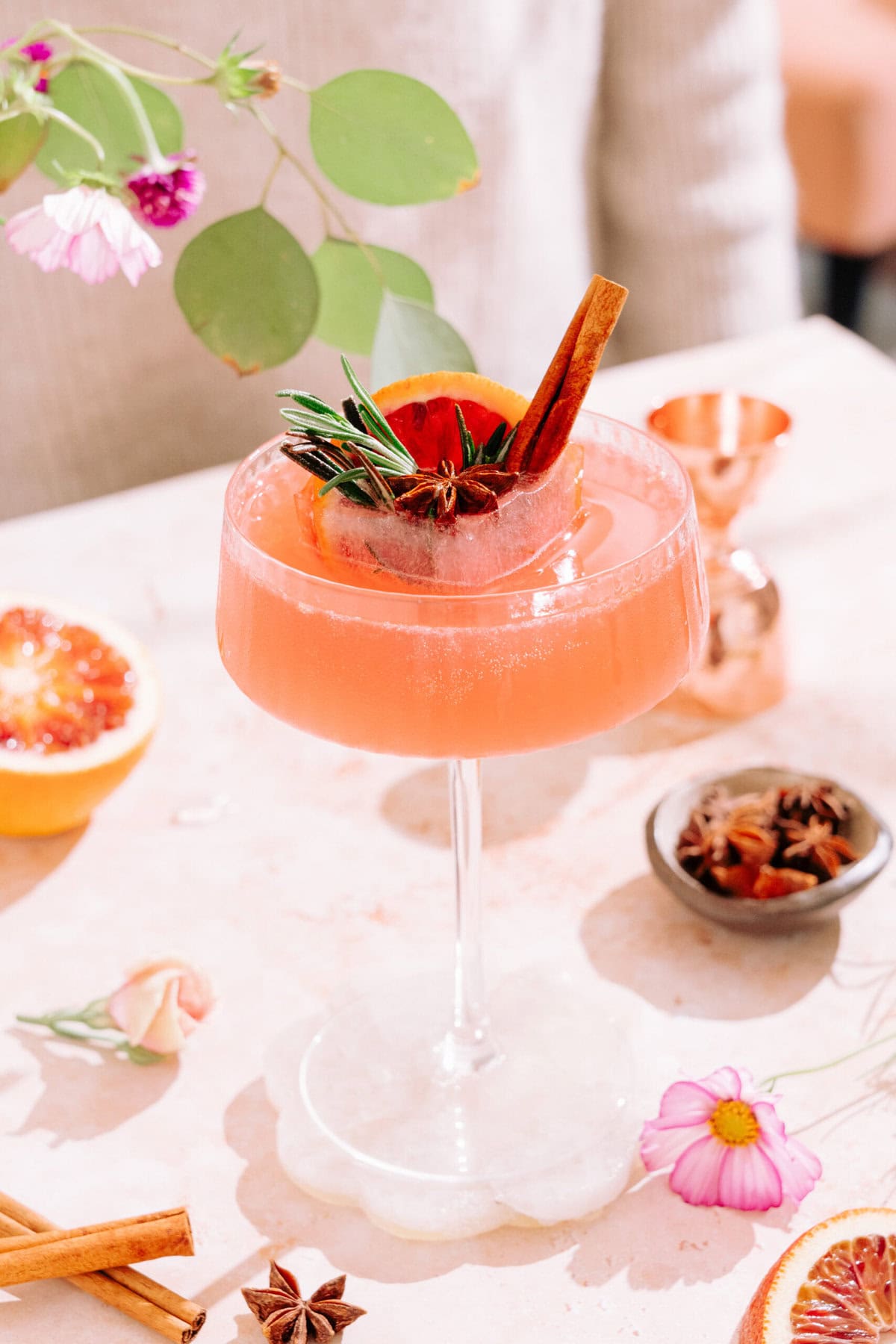 Citrus & Spice Cocktail in coupe glass with festive ice cube surrounded by flowers and spices