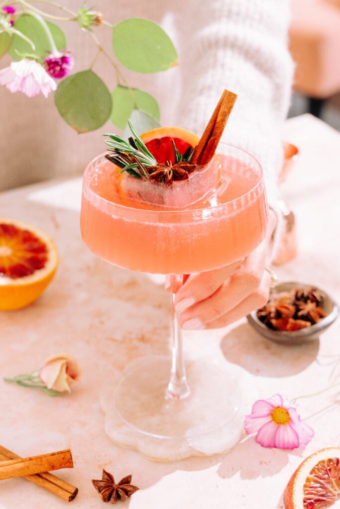 Hand holding Citrus & Spice Cocktail in coupe glass with festive ice cube surrounded by flowers and spices