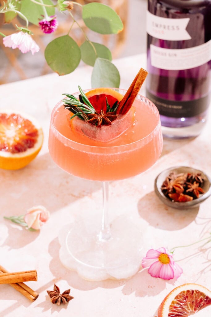 Citrus & Spice Cocktail in coupe glass with festive ice cube surrounded by flowers and spices