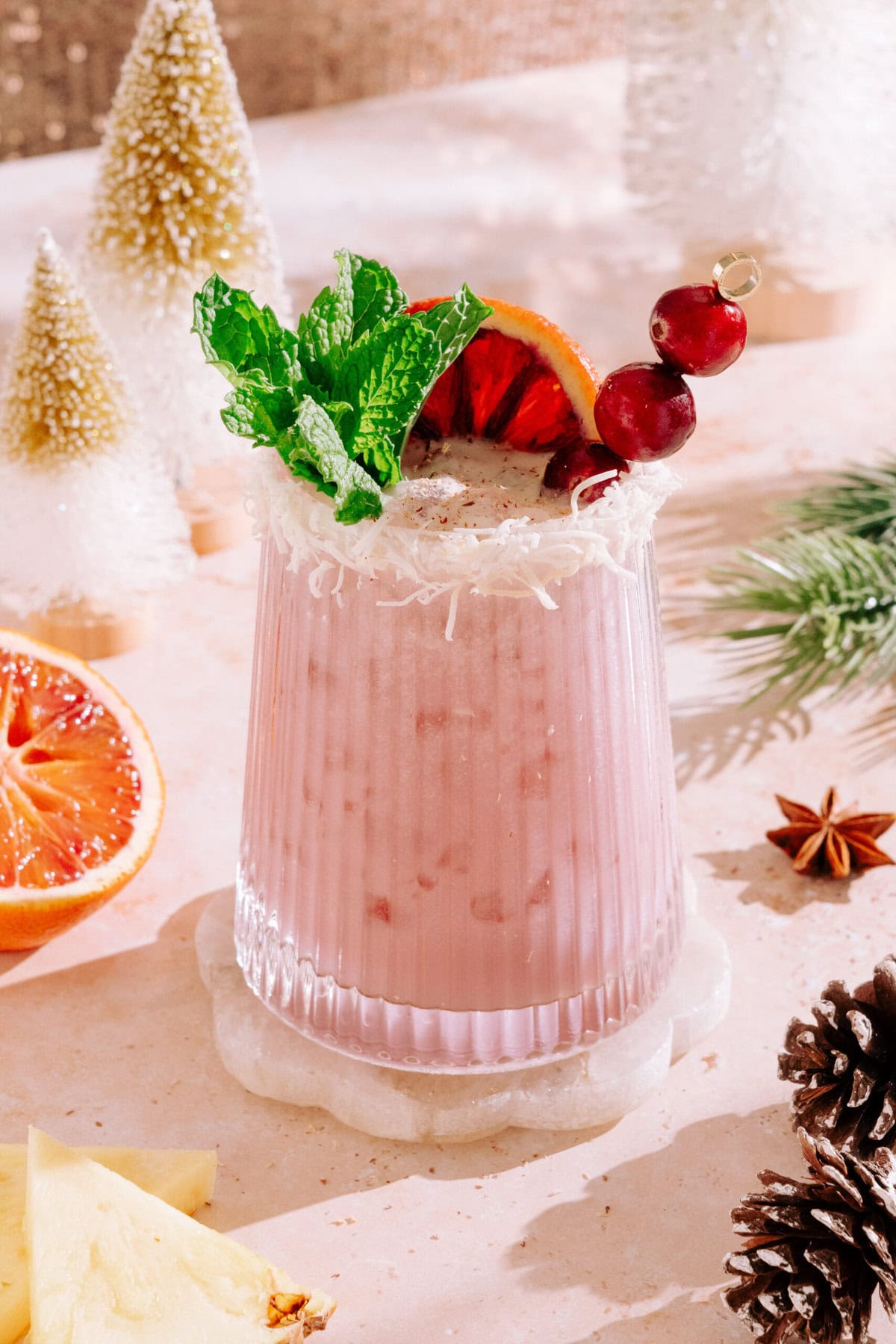 Tequila Painkiller cocktail in small glass decorated with mint, blood orange and cranberries surrounded by Christmas decorations