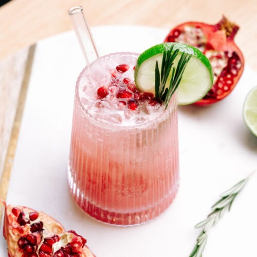 Creamy Pomegranate Mocktail in a short glass garnished with pomegranate seeds, slice of lime, rosemary leaves and salt on the rim of the glass. There is a glass straw in the cocktail.