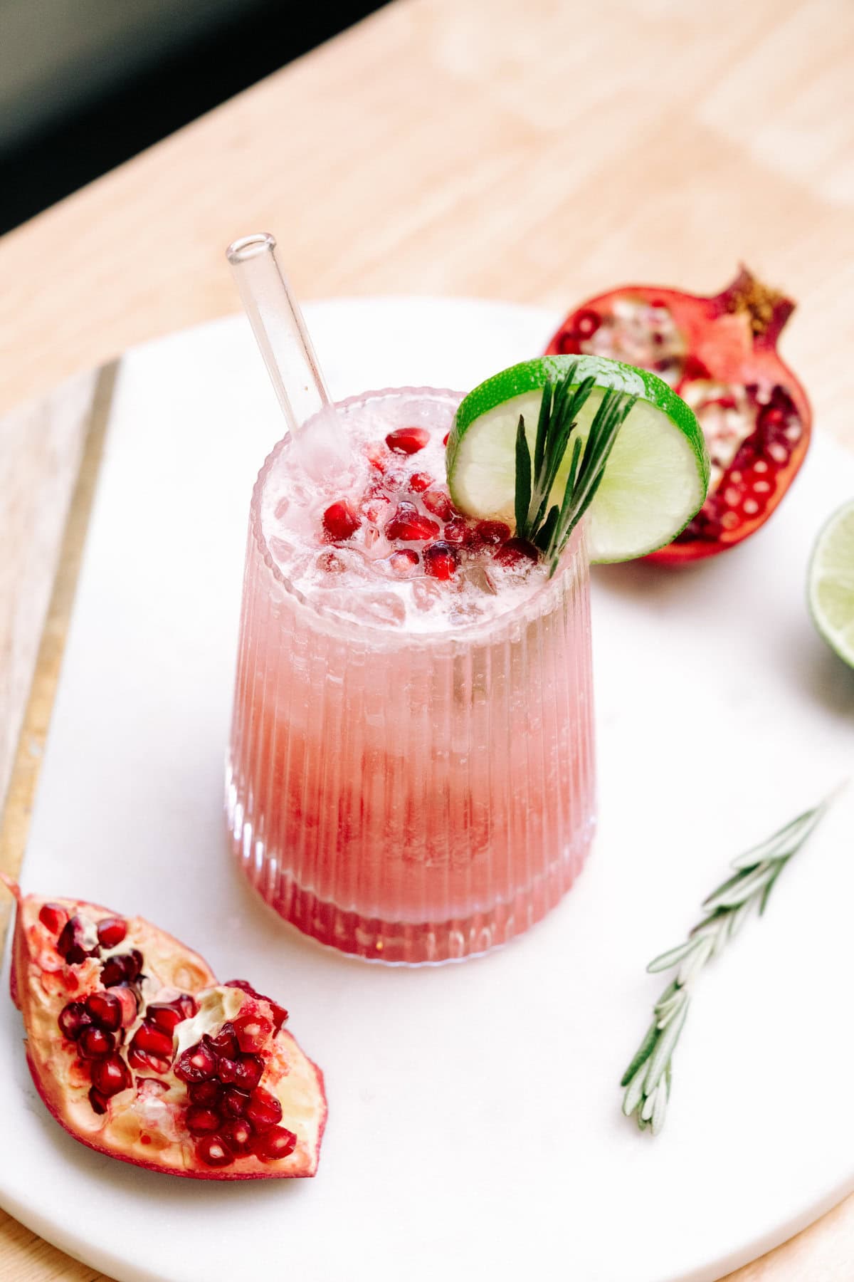 Creamy Pomegranate Mocktail in a short glass garnished with pomegranate seeds, slice of lime, rosemary leaves and salt on the rim of the glass. There is a glass straw in the cocktail.