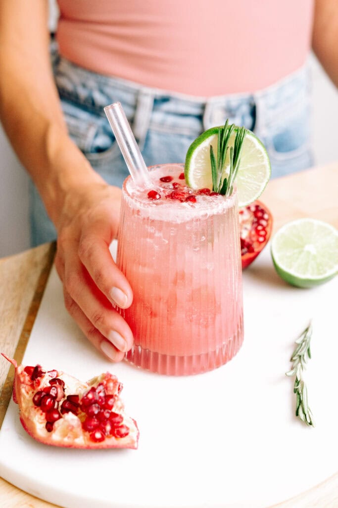 Creamy Pomegranate Mocktail in a short glass garnished with pomegranate seeds, slice of lime, rosemary leaves and salt on the rim of the glass. There is a glass straw in the cocktail and hand holding the glass.