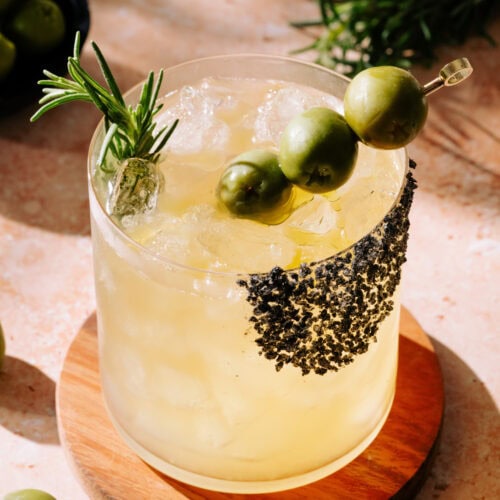 Virgin Dirty Margarita in short glass garnished with 3 olives on a toothpick and rosemary stick.
