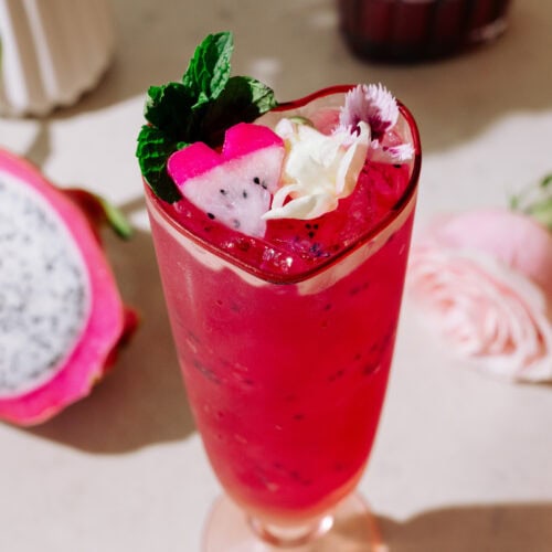 Pretty In Pink cocktail in a high glass with a pink rose and white dragon fruit in the background. The cocktail is garnished with mint leaves, heart-shaped dragon fruit and rose leaves.