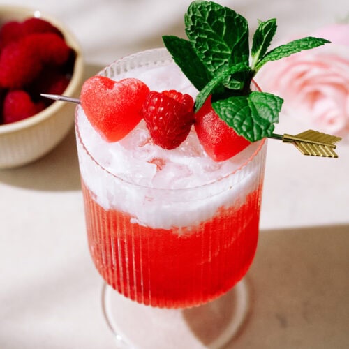 Raspberries & Cream Mocktail in a glass garnished by mint leaves and a toothpick with mix of heart-shaped candy and a raspberry.