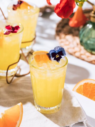 Vibrant yellow cocktail with crushed ice, garnished with an orange slice and edible blue flower, set on a neutral cloth with fresh orange wedges and floral decor.