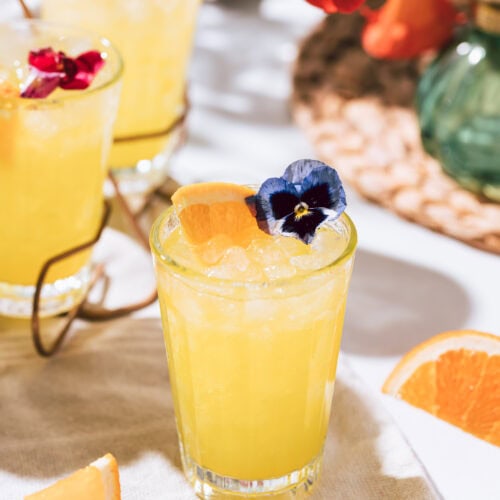 Vibrant yellow cocktail with crushed ice, garnished with an orange slice and edible blue flower, set on a neutral cloth with fresh orange wedges and floral decor.