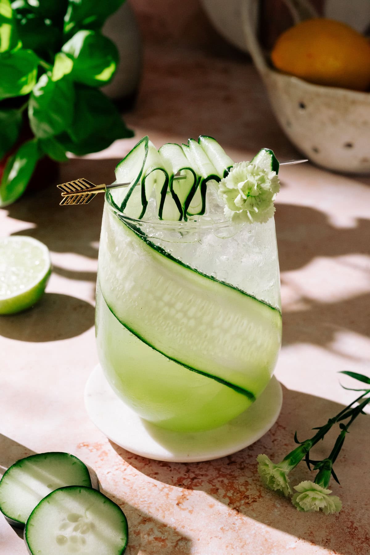 Green dream cocktail in a clear glass with ice, garnished with cucumber slices, a curled cucumber ribbon, and a small white flower on a decorative pick.