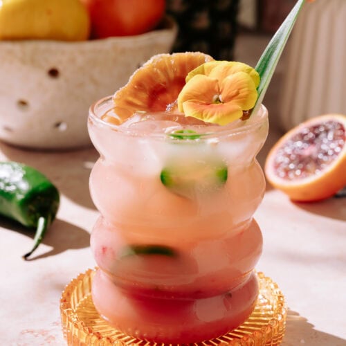 Tropical heat cocktail in a wavy glass with ice, garnished with a dried citrus slice, yellow edible flower, and a pineapple leaf, served on a textured amber coaster.